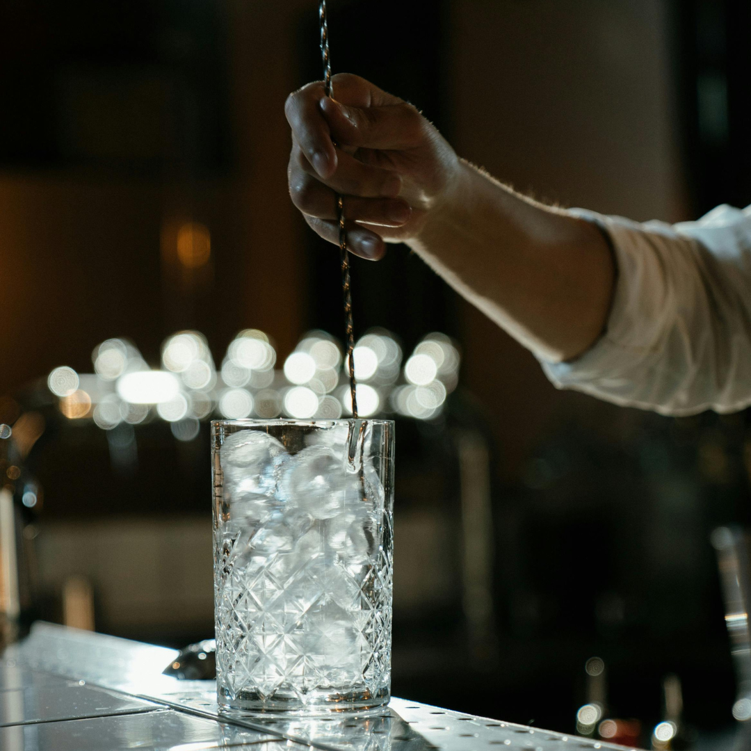 A hand using a bar spoon to stir ice and clear liquid in a clear mixing glass.