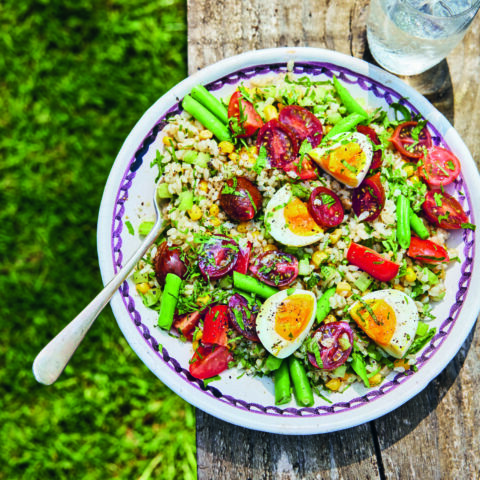 Italian-style rice bowl with asparagus, cherry tomatoes and soft-boiled eggs, served outdoors on a rustic wooden table with a glass of water.