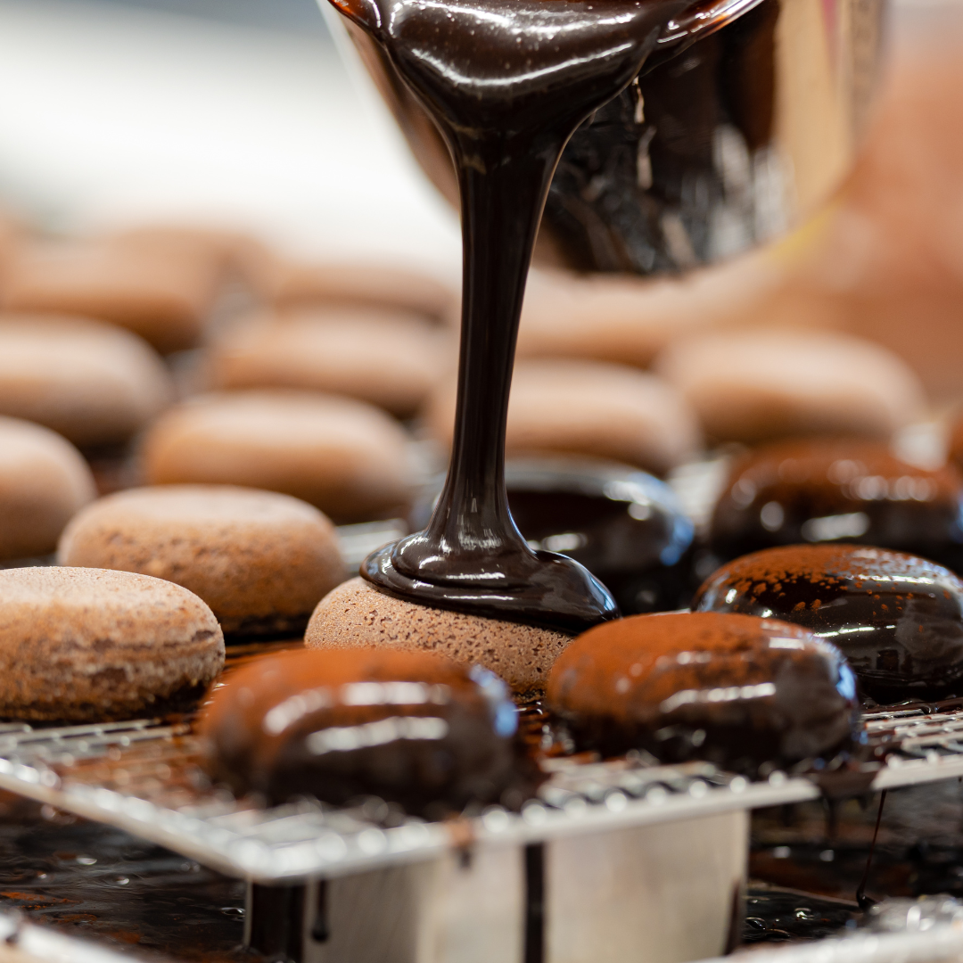 Chocolate ganache being poured over circular chocolate mousse cakes on a wire rack.