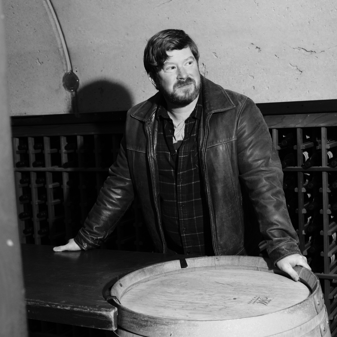 Black and white image of a man in a leather jacket leaning on a wine barrel. 