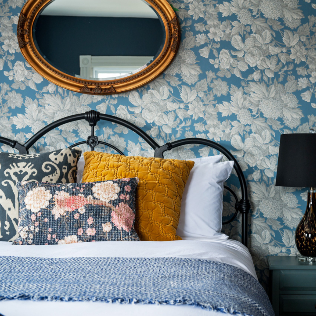 A bed with four pillows against a blue wallpapered wall with a gold mirror.