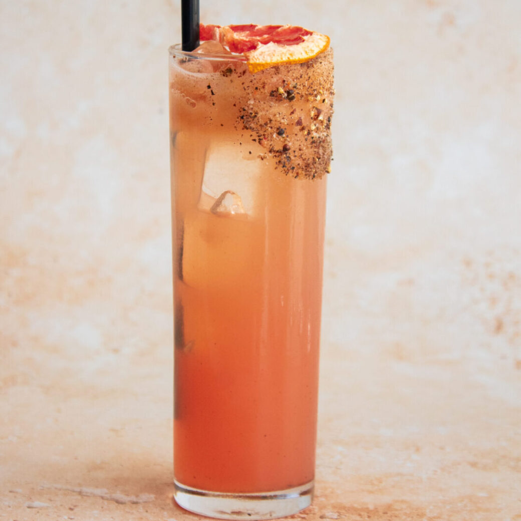 Tall grapefruit cocktail with ice in a Collins glass, garnished with dried citrus and a peppercorn-spiced rim against a soft neutral backdrop.