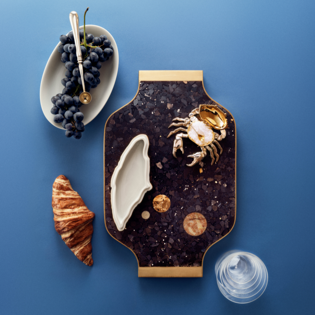 A tablescape featuring a dish with grapes, a croissant and ceramic platter on a blue table. 